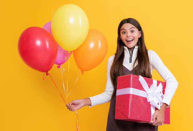Heureux enfant surpris avec des ballons colorés de fête et une boîte cadeau sur fond jaune