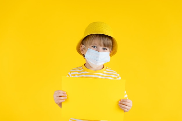 Heureux enfant tenant du papier vierge. Portrait d'enfant portant un masque de protection à l'intérieur