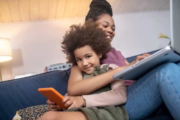 Heureux ensemble. Heureuse maman à la peau foncée travaillant sur un ordinateur portable étreignant une petite fille joyeuse avec une tablette assise sur un canapé à la maison