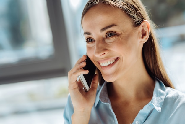 Heureux d'entendre. Femme d'affaires positive attrayante, parler au téléphone intelligent et profiter de la conversation en souriant