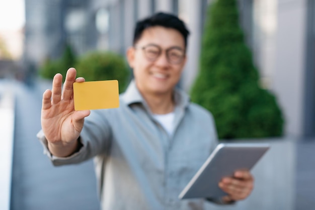 Heureux entrepreneur masculin asiatique tenant une tablette et montrant une carte de crédit à l'appareil photo à l'extérieur mise au point sélective gros plan