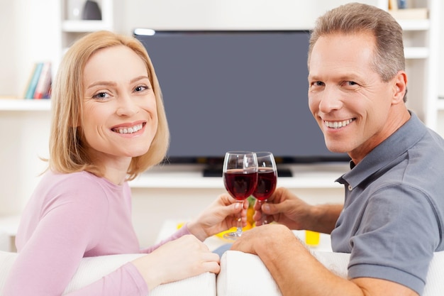 Heureux d'être ensemble. Vue arrière du couple d'âge mûr souriant assis devant la télévision et tenant des verres avec du vin rouge