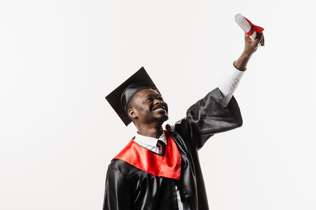 Heureux étudiant africain en robe de graduation noire et casquette lève le diplôme de maîtrise au-dessus de la tête sur fond blanc Un homme africain diplômé est diplômé de l'université et célèbre la réussite scolaire