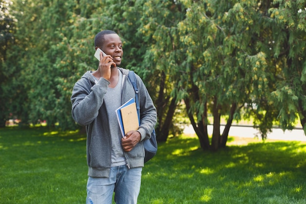 Heureux étudiant afro-américain parlant sur smartphone, tenant des cahiers et un sac à dos derrière ses épaules sur le campus. Technologie, communication, éducation et concept de travail à distance, espace de copie