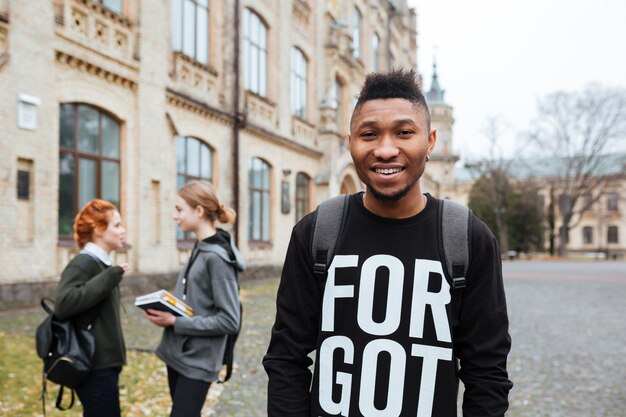 Heureux étudiant afro-américain souriant avec sac à dos debout à l'extérieur sur le campus universitaire