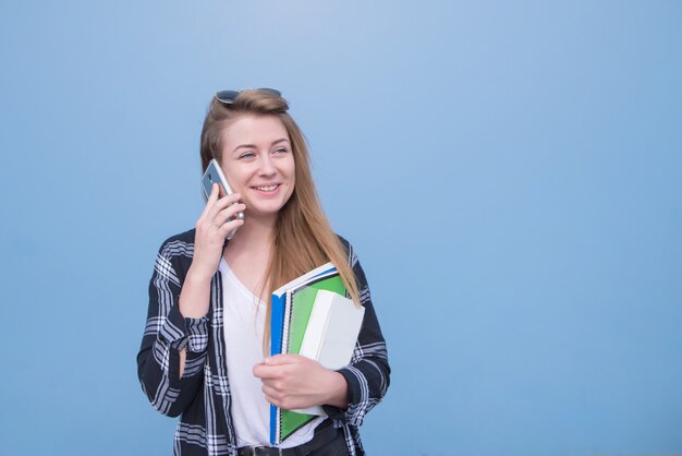 Heureux étudiant parlant sur un smartphone et en détournant les yeux, isolé sur un fond bleu.