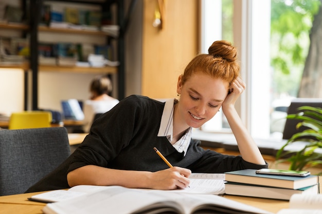 Heureux étudiant Rousse Assis à La Table Avec Des Livres Dans La Bibliothèque, écrire Des Notes