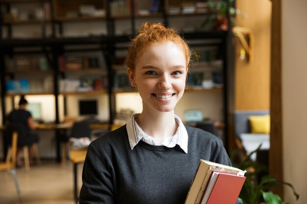 Heureux étudiant rousse posant à l'intérieur dans la bibliothèque tenant des livres