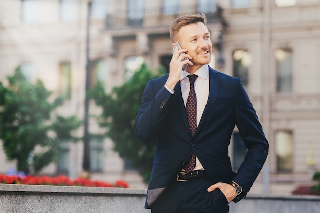 Heureux financier masculin attrayant dans une tenue élégante, à l'aide d'un smartphone moderne