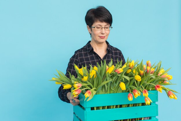 Heureux fleuriste femme d'âge moyen portant des lunettes avec boîte de tulipes sur bleu.