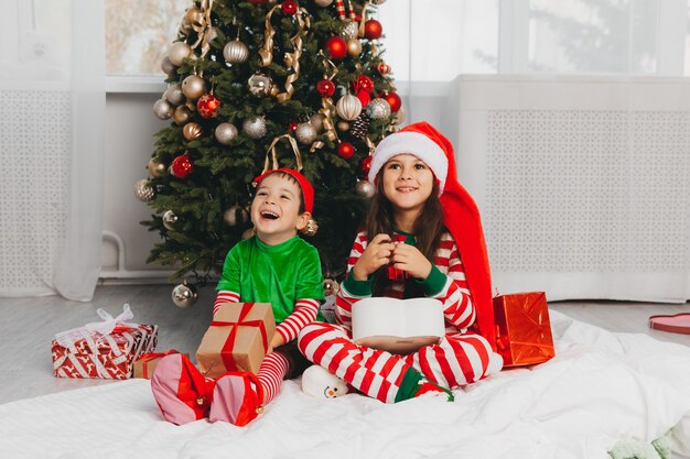 Heureux frère et sœur déguisés en Père Noël sont assis près de l'arbre de Noël à la maison dans le salon avec des cadeaux. Noël