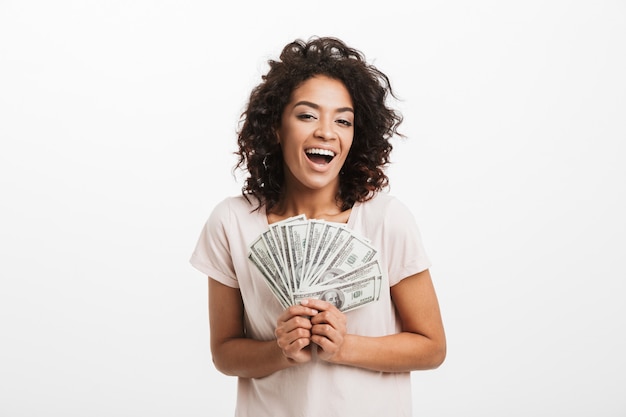 Heureux gagnant femme américaine avec une coiffure afro et grand sourire tenant de l'argent en espèces, isolé sur un mur blanc