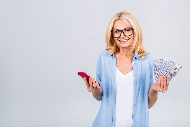 Heureux gagnant ! Image d'une femme senior heureuse mature isolée sur un mur de fond blanc regardant la caméra tenant de l'argent à l'aide d'un téléphone portable