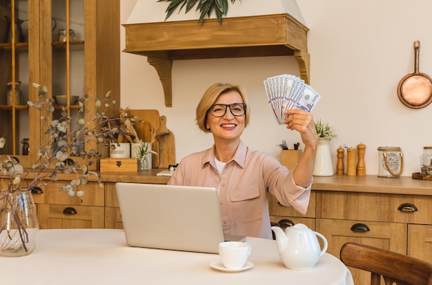 Heureux gagnant. Joyeuse femme âgée souriante debout dans la cuisine et utilisant son ordinateur portable et ses factures d'argent, pigiste travaillant à la maison.