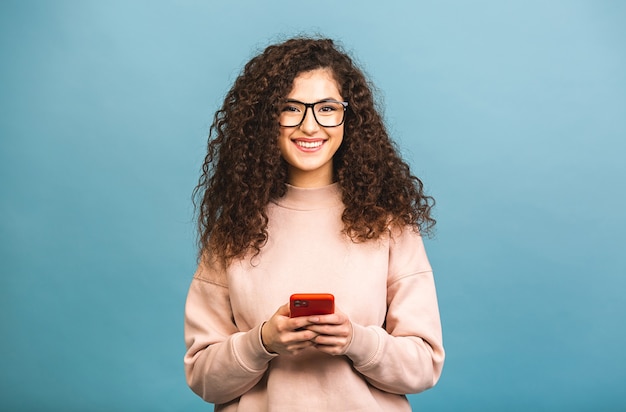 Heureux gagnant! Portrait de femme étudiante bouclée décontractée surpris surpris souriant tenant un smartphone isolé sur fond bleu.