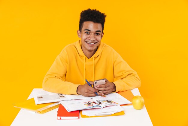 Heureux garçon adolescent étudiant assis au bureau avec des manuels isolés sur un mur jaune, à l'aide d'un téléphone portable