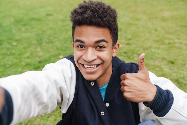 Heureux garçon adolescent prenant un selfie en se tenant debout sur une pelouse, montrant les pouces vers le haut