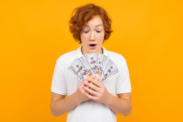 Heureux garçon adolescent surpris en t-shirt blanc tenant un fan de billets d'argent dans ses mains sur jaune