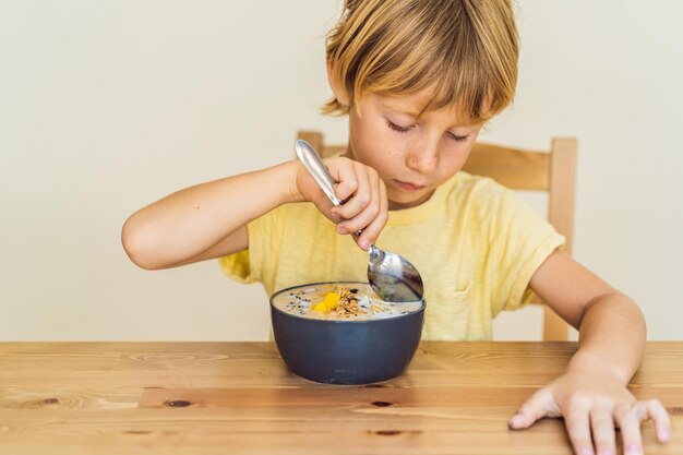 Heureux garçon assis et mangeant un bol de smoothie frais avec du granola aux fruits du dragon à la mangue et des graines de chia