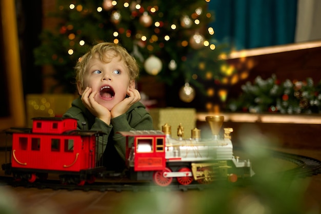 Heureux garçon aux cheveux bouclés joue avec un train jouet magique à l'arbre de Noël