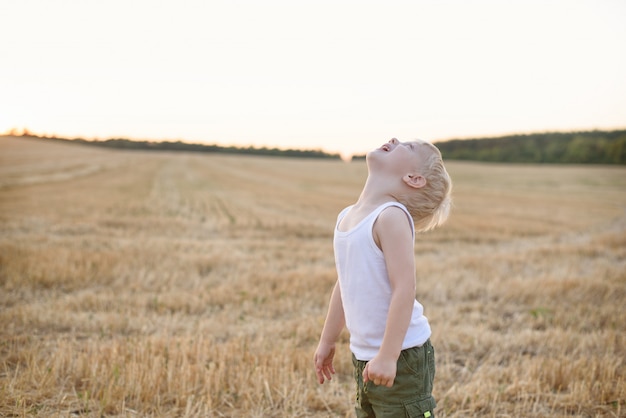 Heureux garçon blond se dresse la tête sur un champ de blé tondu. Coucher du soleil