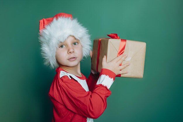 Heureux garçon caucasien souriant en santa tee shirt et chapeau tenant une boîte avec un cadeau de noël