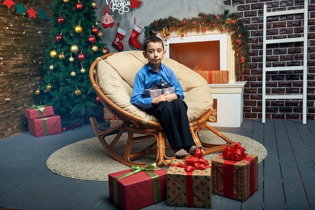 Heureux garçon dans une chaise confortable près de l'arbre de Noël près de la cheminée avec beaucoup de cadeaux.