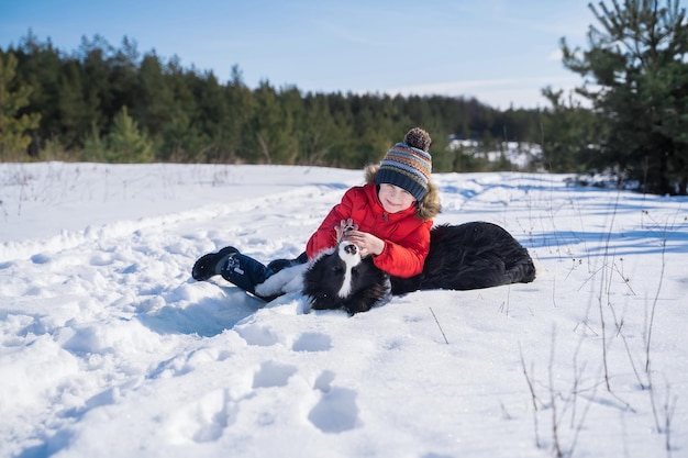 Heureux garçon dans une veste rouge jouant avec un border collie à l'extérieur sur fond d'hiver