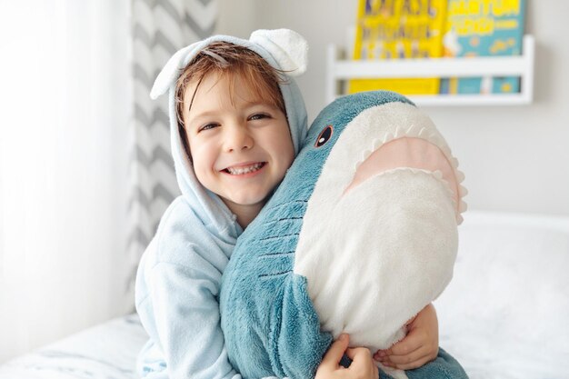 Heureux garçon enfant riant en peignoir bleu doux après le bain jouer avec un jouet de requin sur un lit blanc dans une chambre ensoleillée Concept de lavage et d'hygiène pour enfants