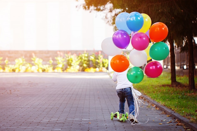 Heureux garçon jouant avec un bouquet de ballons à l&#39;extérieur et sur un scooter.