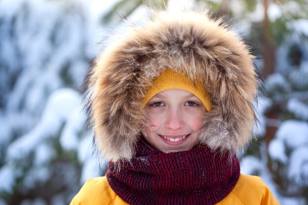 Heureux garçon mignon dans un chapeau, une écharpe et une capuche sur la tête souriant dans le parc d'hiver pendant les chutes de neige. Portrait d'hiver