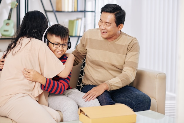 Heureux garçon préadolescent vietnamien dans de nouveaux écouteurs serrant ses parents après les avoir reçus comme cadeau d'anniversaire