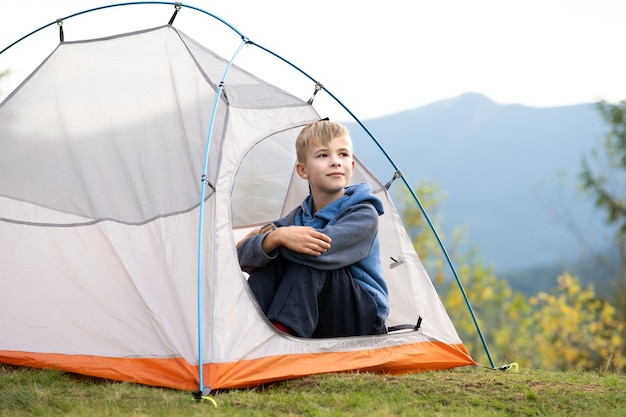 Heureux garçon se reposant seul dans une tente touristique au camping de montagne, profitant d'une vue sur la belle nature estivale. Concept de randonnée et de mode de vie actif.