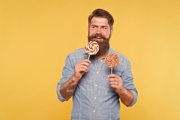 Heureux hipster barbu manger de savoureux bonbons tourbillon arc-en-ciel apparaît fond jaune délicieux