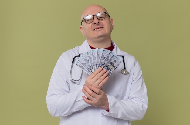 Heureux homme adulte avec des lunettes en uniforme de médecin avec stéthoscope tenant de l'argent