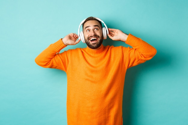 Heureux homme adulte en pull orange, regardant et écoutant de la musique dans des écouteurs, debout sur fond bleu.