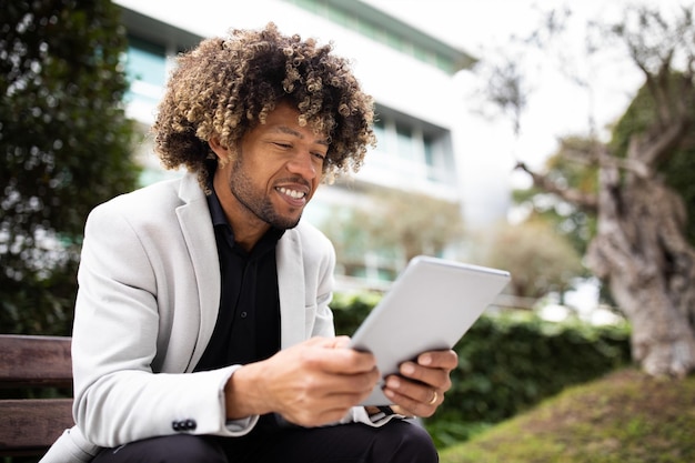 Photo heureux homme d'affaires d'âge moyen noir confiant en costume à l'aide d'une tablette numérique assis sur un banc près de