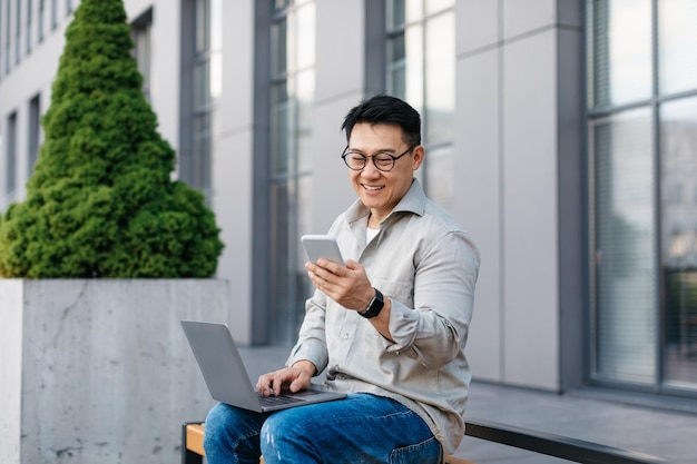 Heureux homme d'affaires asiatique d'âge moyen envoyant des SMS sur un téléphone portable et utilisant un ordinateur portable discutant avec le client à l'extérieur