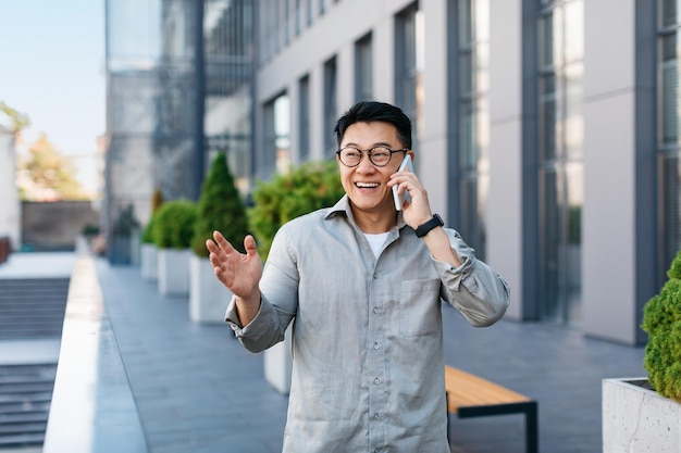 Heureux homme d'affaires asiatique prospère parlant sur smartphone debout à l'extérieur contre l'espace libre du centre de bureau moderne