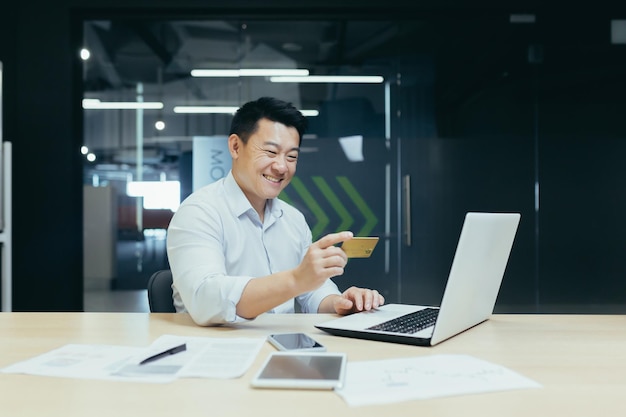 Heureux homme d'affaires asiatique travaillant dans un bureau moderne faisant un transfert d'argent en ligne à l'aide d'un ordinateur portable et