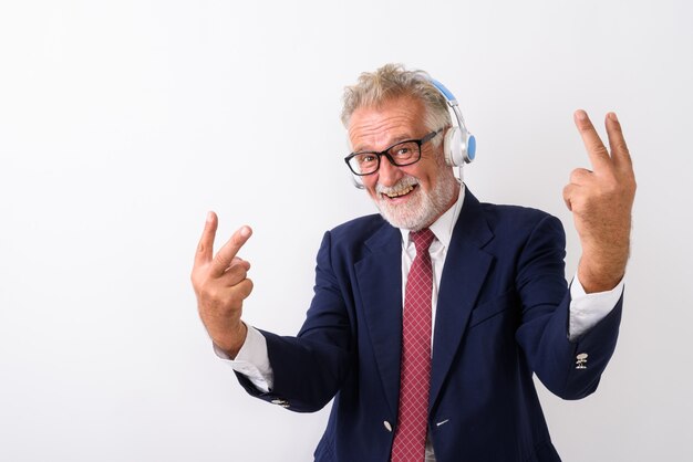 Heureux homme d'affaires barbu senior souriant et posant avec des signes de paix tout en écoutant de la musique sur blanc