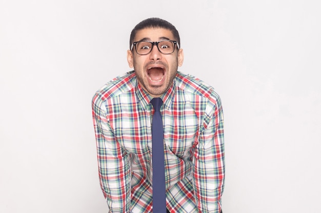 Heureux homme d'affaires barbu surpris en chemise à carreaux colorée, cravate bleue et lunettes noires debout et regardant la caméra avec le visage étonné et la bouche ouverte. tourné en studio, isolé sur fond gris