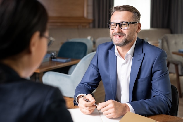 Heureux homme d'affaires confiant en tenues de soirée et lunettes regardant un collègue ou un client tout en lui parlant lors d'une réunion au restaurant