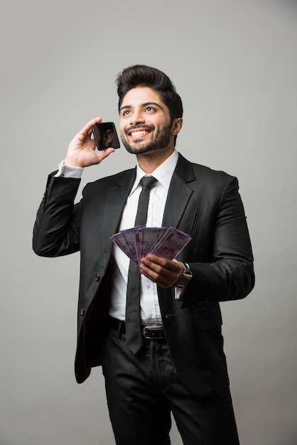 Heureux homme d'affaires indien barbu tenant un ventilateur d'argent à la main tout en parlant au téléphone, debout isolé sur fond gris