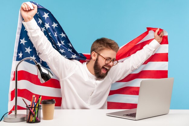 Photo heureux homme d'affaires levant le drapeau américain, regardant un programme patriotique en direct sur un ordinateur portable sur le lieu de travail et criant de joie célébrant le jour de l'indépendance des états-unis le 4 juillet, des nouvelles sur le soutien aux entreprises du gouvernement