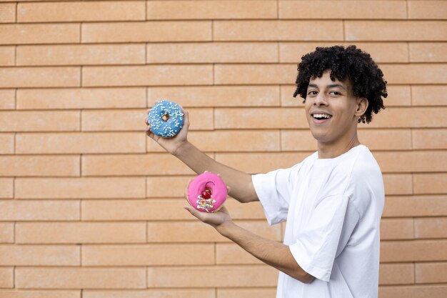 Heureux homme africain souriant debout sur le côté portant un t-shirt blanc tenant des beignets roses et bleus