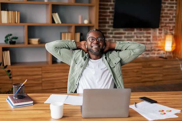 Heureux homme afro-américain d'âge moyen réussi dans des lunettes et décontracté dans des écouteurs sans fil se reposant du travail