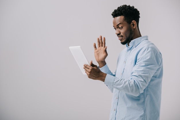 Heureux homme afro-américain agitant la main tout en tenant une tablette numérique isolée sur gris