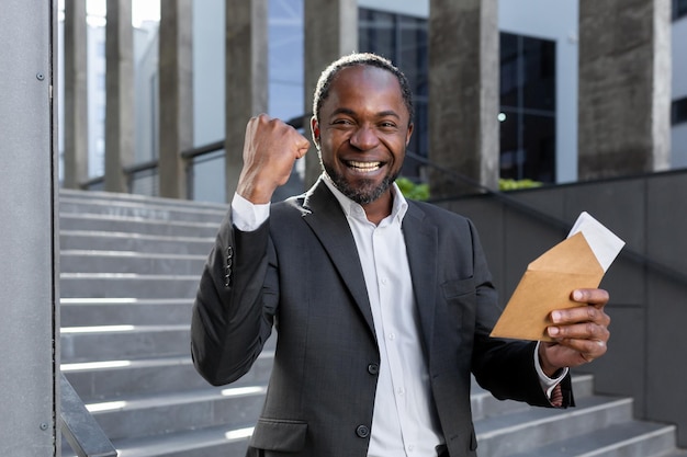 Heureux homme afro-américain debout à l'extérieur près du centre de bureau en costume et tenant une enveloppe avec