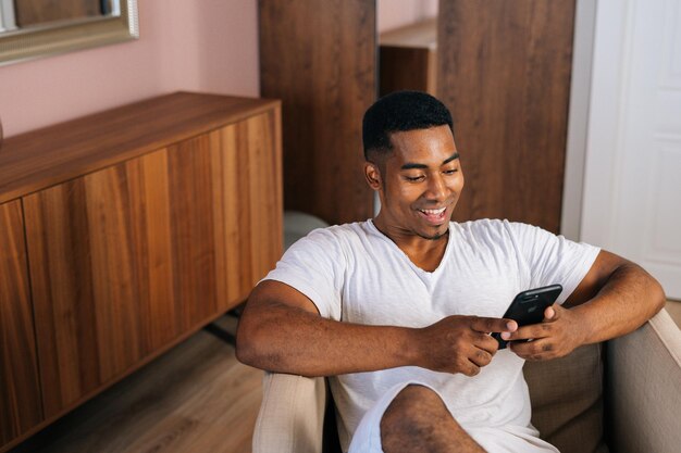 Heureux homme afro-américain souriant en T-shirt blanc assis dans un fauteuil dans un salon confortable et lumineux et utilisant un téléphone portable. Beau mâle naviguant sur Internet sur téléphone portable. Travelling au ralenti.